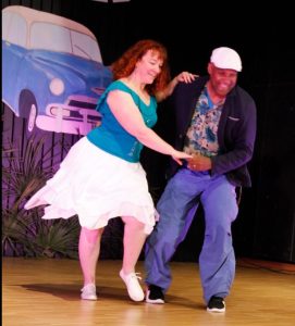 Un couple danse sur une scène, avec une voiture bleue en arrière-plan.